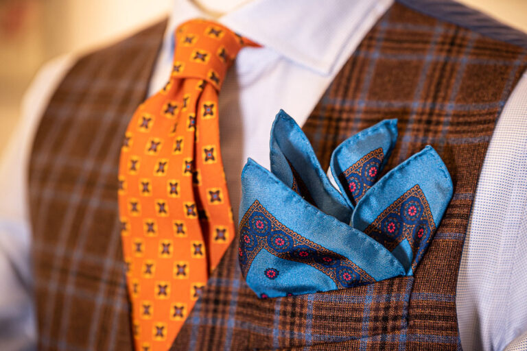 Dettaglio outfit invernale con gilet Principe di Galles marrone e azzurro, cravatta arancione e fazzoletto azzurro disegno cachemire in seta